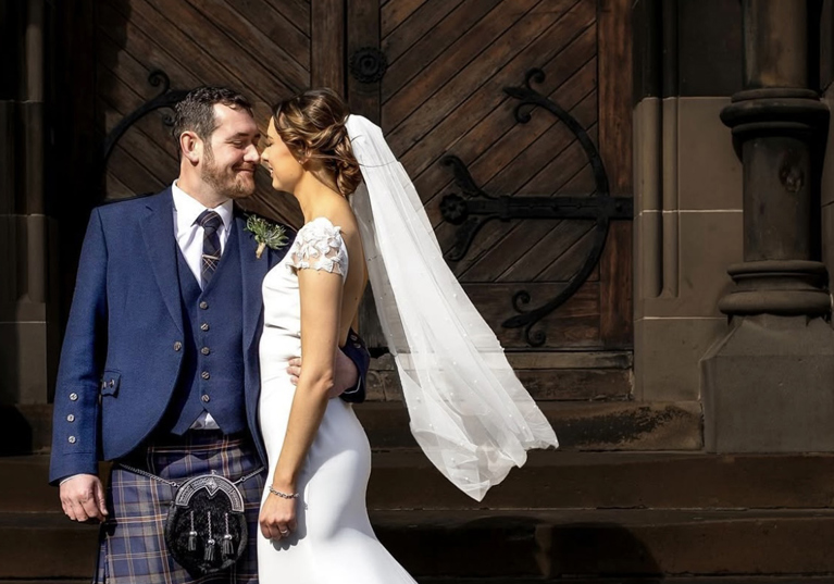 Bride wearing veil and groom wearing a kilt standing outside venue in an embrace while smiling at each other